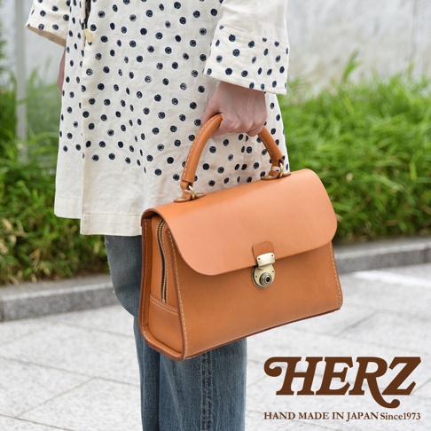 日本HERZ手工皮革多功能收納單肩包，焦糖棕色，側面拉鍊設計，正面鎖扣，品牌標誌印製在右下角。