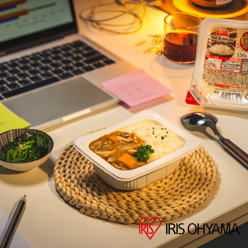 一盒白飯配咖哩，內有紅蘿蔔塊、香菇片及綠色花椰菜，飯上撒有黑芝麻，置於白色塑膠餐盒內，餐盒放置於草編墊上。