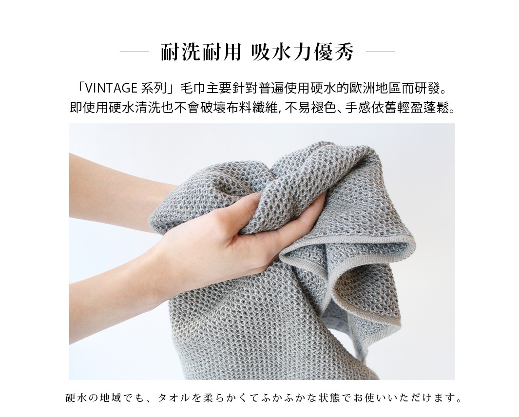 一雙手拿著灰色鬆餅紋棉麻混紡大浴巾，浴巾質地柔軟，邊緣有細緻的包邊處理。