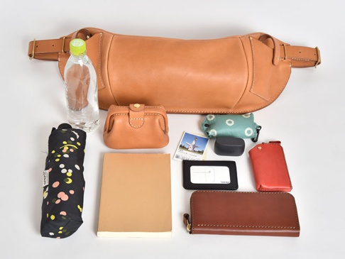 職人手作皮革大容量斜背包，淺棕色皮革材質，搭配金色金屬扣環。包內物品包含：一瓶礦泉水、黑色圓點圖案摺疊傘、淺棕色筆記本、紅色皮革卡夾、黑色長夾、黑色無線耳機盒、淺綠色印花收納包及一張票券。