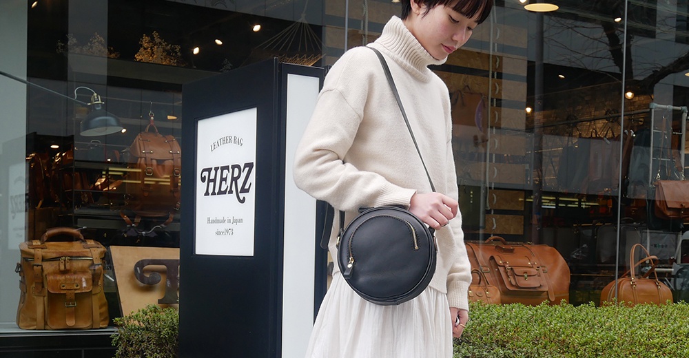 穿著米色高領毛衣的女士，身揹一個黑色皮革小圓包，包包圓潤，正面有金色拉鍊裝飾，整體造型簡潔大方。