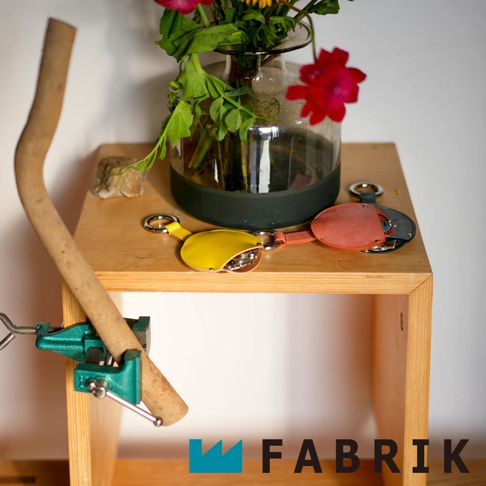 FABRIK 品牌標誌，旁邊擺放著黃色、珊瑚粉色和深灰色的皮革圓形收納鑰匙圈，架上還有一個花瓶裡插著鮮花，左邊有一個綠色的鉗子夾著一根木棍。