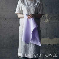 一位身穿淺色圍裙的人手持一條淡紫色、觸感柔軟的毛圈浴巾，浴巾材質看起來吸水性佳。