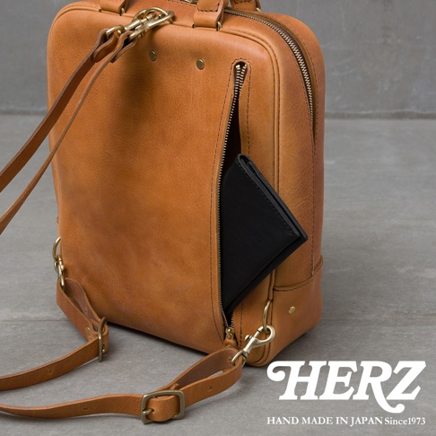 一個棕色皮革後背包，拉鍊口袋裡露出一個黑色皮夾，包包上有可拆卸的肩帶和金屬配件，右下角有HERZ品牌標誌和HAND MADE IN JAPAN Since1973文字。