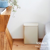 米色方形垃圾桶，帶有掀蓋，置於木質地板上，靠近木製桌子和白色牆壁。