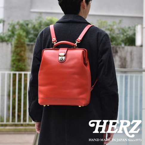 一位背著日本HERZ職人手作皮革後背包的男子，背包呈現搶眼的紅色調，採用獨特的圓釦設計與硬挺箱型結構，上方設有提把，並搭配金屬扣環與底部金屬腳釘，展現精緻的皮革手工質感。