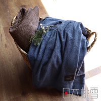 編織籃中擺放著一深棕色與一深藍色的超長棉毛巾被，質地柔軟，深藍色毛巾被上裝飾有幾枝松枝，毛巾被的標籤清晰可見，顯示品牌名稱。