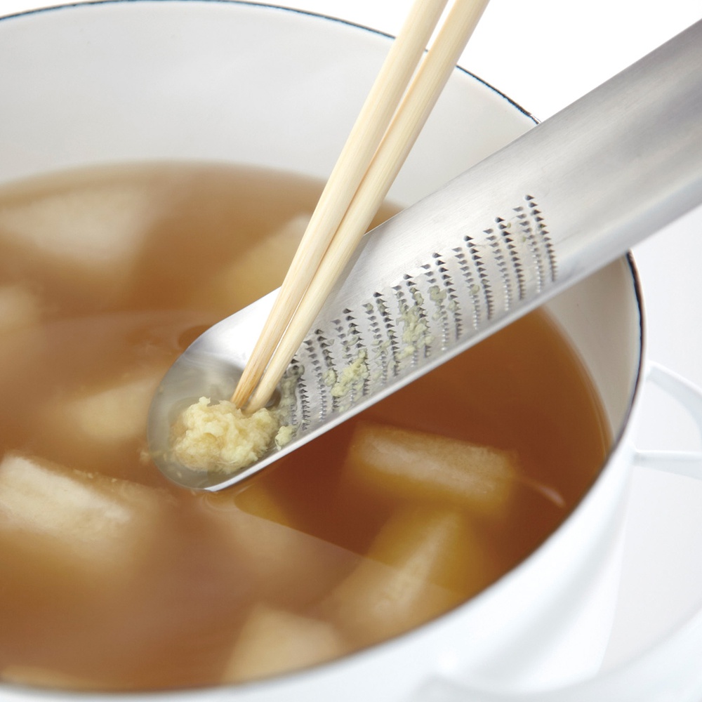 日製不鏽鋼磨泥器，帶有細緻的磨齒，一端為湯匙形狀，盛有磨碎的薑末，另一端置於裝有燉湯的白色鍋中，湯中有塊狀食材，由一雙木筷夾著磨泥器。