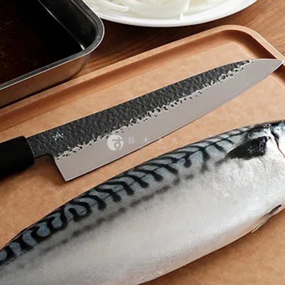 玄 / Chef's knife 主廚刀，18公分，銀色鋼製刀刃，黑色刀柄，刀刃表面有槌目紋理，旁邊擺放著一條正在處理的鯖魚。