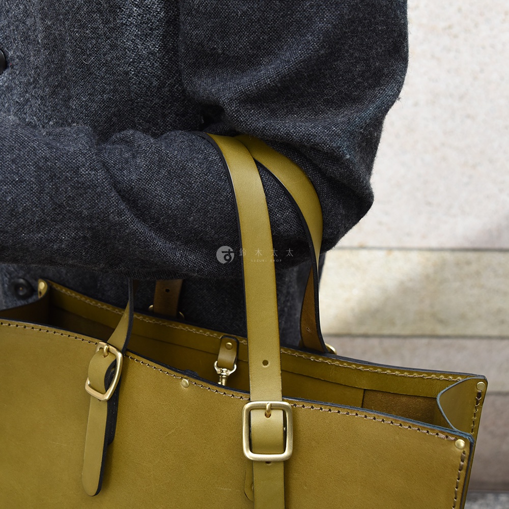 橄欖綠皮革托特包，附有可調節的皮質提把和金色金屬扣。包身線條簡潔，皮革邊緣有縫線細節，展現職人手作質感。