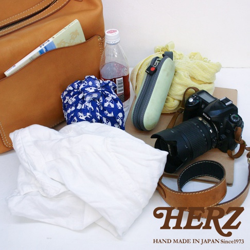 一個棕色皮革厚實單肩波士頓包，部分敞開，露出白色衣物、藍白圖案束口袋、一瓶飲料、一個淺綠色筆袋、一條黃色絲巾、一台黑色 Nikon 相機及鏡頭，以及一條棕色皮革背帶。