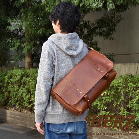 一個男人揹著棕色皮革復古郵差包，包包有翻蓋設計，金屬鉚釘點綴，在戶外環境中展示。