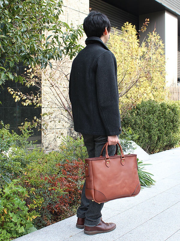 男性手持一個棕色皮革托特包，包包有兩個提把、側邊扣環和金屬腳釘。