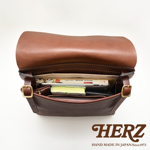 職人手作皮革經典郵差包，深棕色，內部裝有書籍、平板電腦和筆記本，展現皮革材質的質感與實用性。