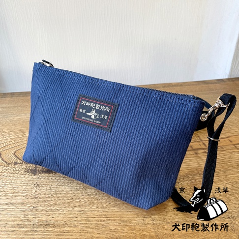 深藍色刺子織隨身斜背包，帶有黑色織帶和金屬掛鉤，包面上方有一個印有「犬印鞄製作所」字樣和圖案的黑色標籤。