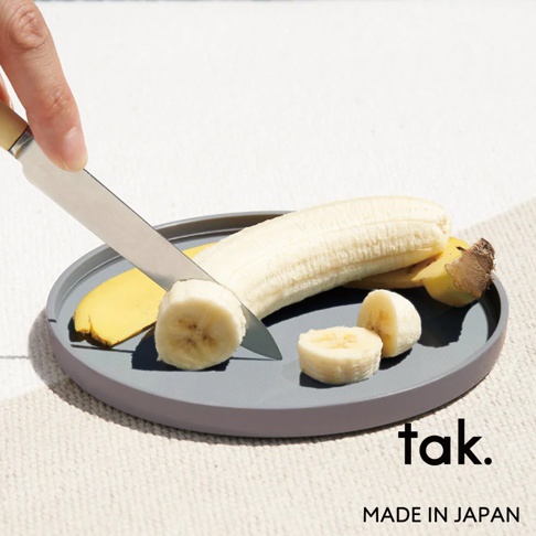 灰色圓形餐盤，上面放著一根剝皮的香蕉，以及切片的香蕉。一把刀正切著香蕉。