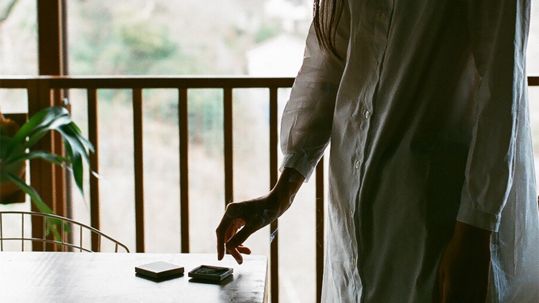 桌面上擺放著一組香氛火柴禮盒，其中兩個方形深色盒裝產品，一個盒蓋已打開，露出內部淺色火柴，旁有一人手部伸出，似乎正要取用火柴，火柴燃燒後產生淡淡煙霧。