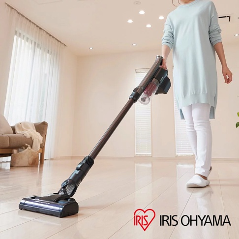 淺藍色長版上衣與白色長褲的女子，手持深棕色金屬桿、黑色吸頭、透明集塵筒的氣旋式超輕量無線手持吸塵器，正在淺色木質地板上進行清潔。