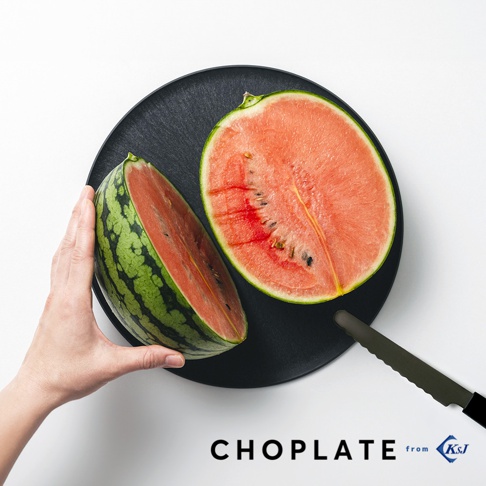 黑色圓形兩用砧板盤上放著切半的西瓜，一隻手扶著其中一半，另一半則放在盤中。盤旁有一把黑色鋸齒刀。
