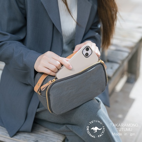 一名女子身穿灰色西裝外套，手拿著手機，正將其放入一個灰色的小包中，包上有黃銅色拉鍊和皮革裝飾，包上印有TAKARA-MONO TUTUMU Made in jpn 的字樣
