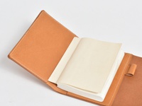 職人手作皮革A6記事本書套，淺棕色皮革，內有白色紙張的記事本，書套邊緣有白色縫線，並設有皮革筆套。