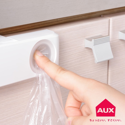 白色的塑膠收納袋架，架上有個圓形開口，手指正將一個透明塑膠袋從開口抽出，架子固定在淺木紋的櫃體上。