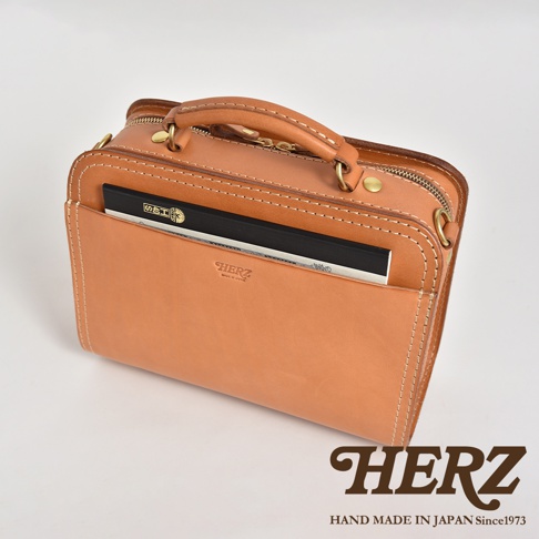 職人手作皮革兩用方形單肩包，以自然色皮革製成，搭配白色縫線。包包設有頂部提把、拉鍊開口，前方有一個插袋，內含筆記本。