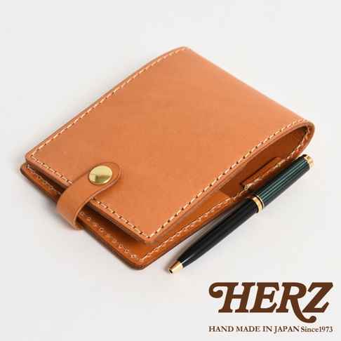 職人手作皮革便利貼收納套，淺棕色皮革，附有金色按扣，搭配一支綠色條紋的黑色鋼筆。