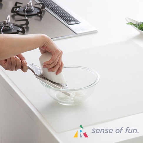 一張白色耐高溫料理台保護墊，上面有一雙手正在使用刨絲器將白蘿蔔刨成絲，刨絲器置於透明玻璃碗上方。