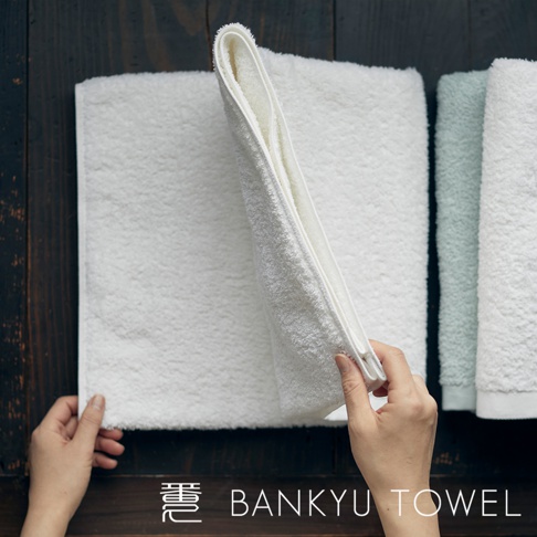 雙手展開一條白色長纖維毛巾，展示其柔軟蓬鬆的質地，旁邊擺放著一條淺綠色和另一條白色的毛巾。