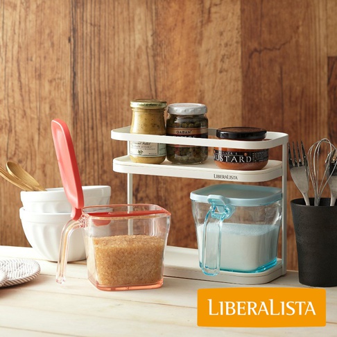 LIBERALISTA 調味料罐收納架上擺放著多個玻璃罐，其中一個罐子上印有「MUSTARD」字樣。右側有一個黑色杯子，裡面插著幾個金屬打蛋器。前景是一個透明的塑膠量杯，裡面裝有褐色糖粒，旁邊還有兩個白色碗和木質湯匙。