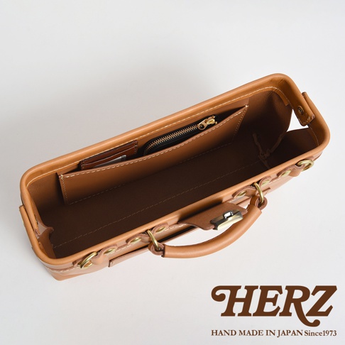 HERZ 品牌標誌與「HAND MADE IN JAPAN Since1973」字樣，搭配一個傾斜拍攝的棕色皮革手提包內部，展示了其隔層和拉鍊小袋。