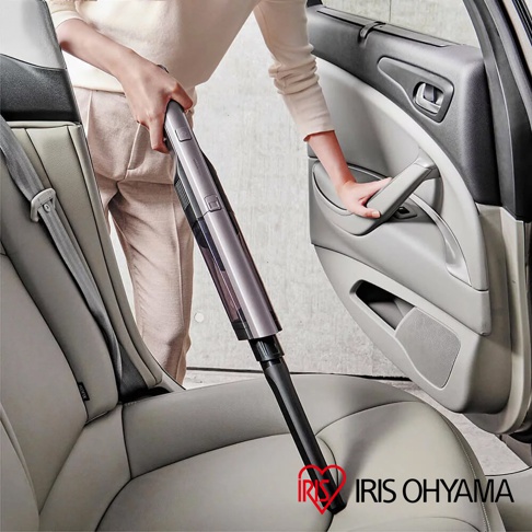 灰色金屬與透明塑膠材質的充電式輕巧吸塵器，正用於清潔汽車座椅。吸塵器主體為灰色，集塵筒為透明設計，可見內部。
