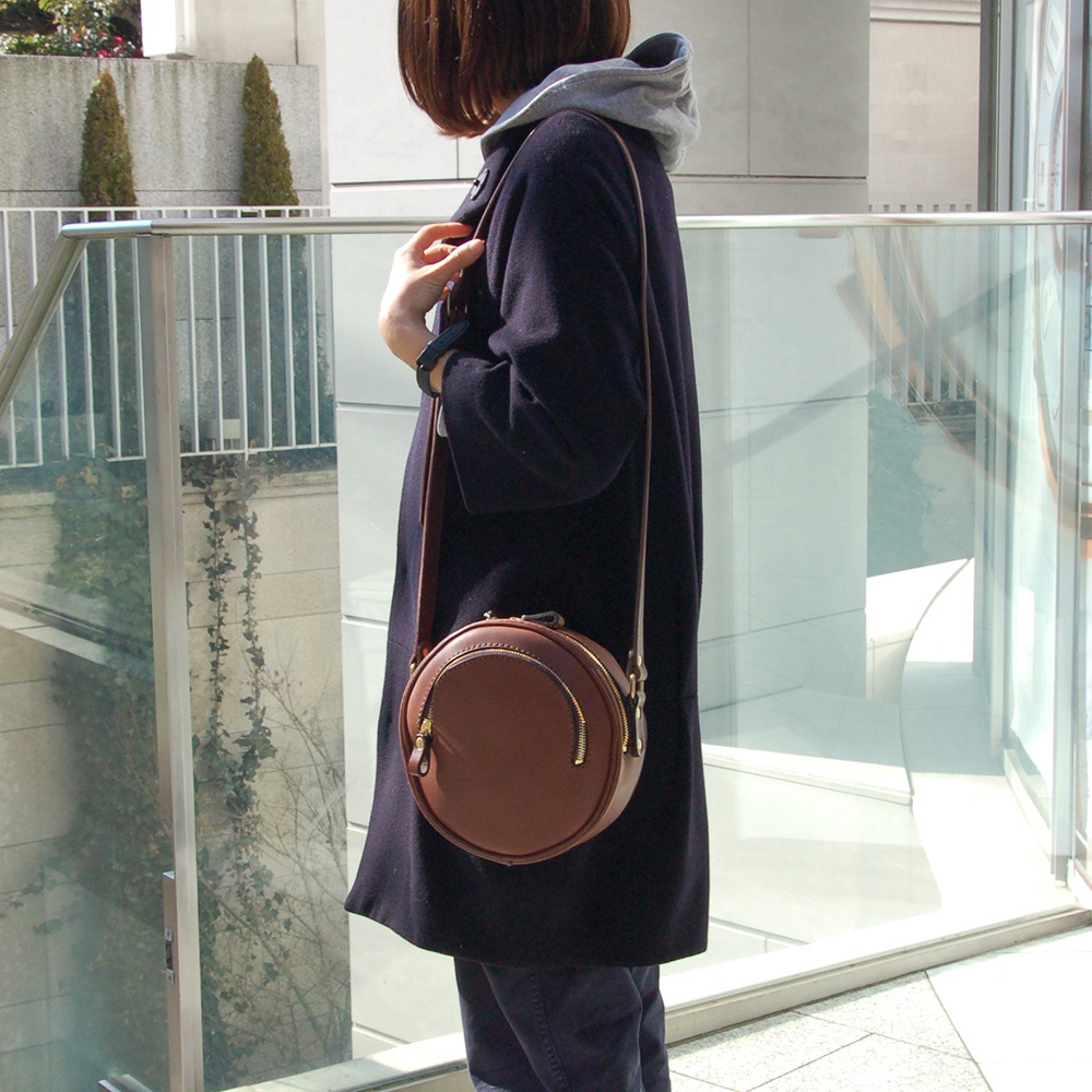 職人手作皮革小圓包，咖啡色皮革，圓形包身，正面有拉鍊開口，搭配長皮質背帶。