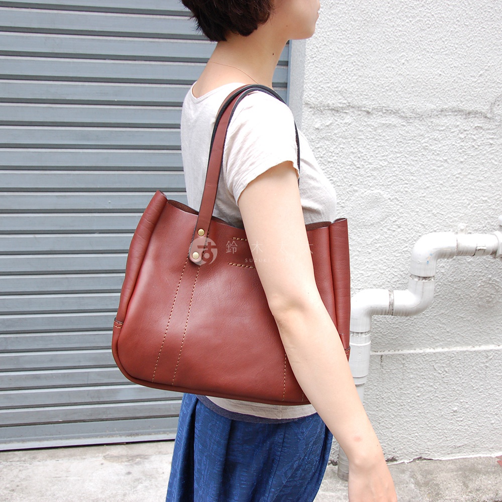 女性側身展示一個紅棕色皮革托特包，包包設計簡約，配有兩條皮革提把，包身有明顯的縫線細節。