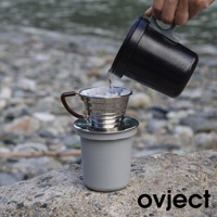 一個銀色金屬濾杯，帶有棕色把手，置於灰色杯子上。一個黑色水壺正將熱水倒入濾杯，濾杯內有咖啡濾紙。