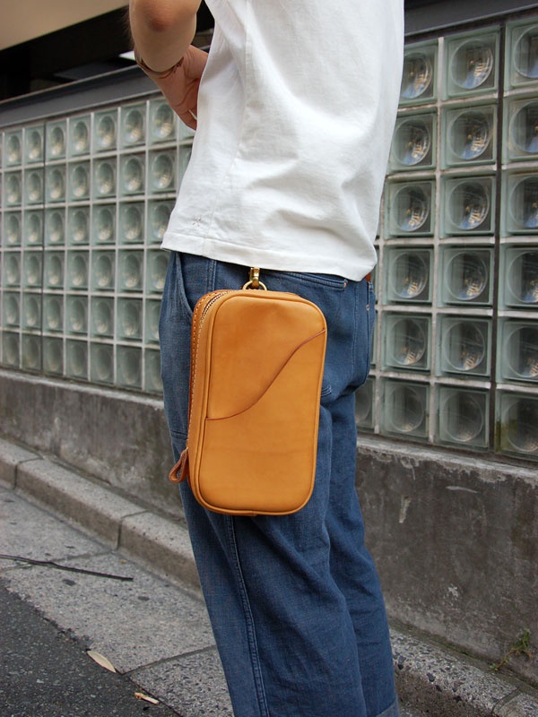 一款淺棕色皮革製作的直立型斜背包，附有拉鍊開合和側邊縫線細節，透過金色D型環懸掛於牛仔褲腰帶上。