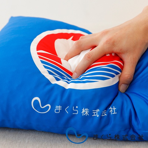 藍色布面小抱枕，印有白色愛心圖案與日文「まくら株式会社」字樣，抱枕中央有紅色與藍白色條紋的圓形標誌，手正按壓抱枕，展現其柔軟材質。