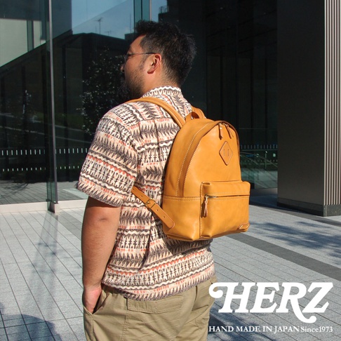 一名男性背著一個黃褐色皮革後背包，背包上有一個拉鍊前袋和一個方形商標。