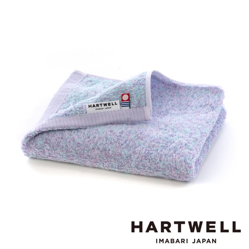 這是一條摺疊起來的Hartwell品牌今治毛巾，材質呈現淡雅的藍紫色與粉色混紡色調，毛巾表面具有細膩且柔軟的無撚棉質紋理，邊緣處縫有標示著Imabari Japan與品牌名稱的紅色圓形商標標籤，整體外觀呈現清新舒適的質感。