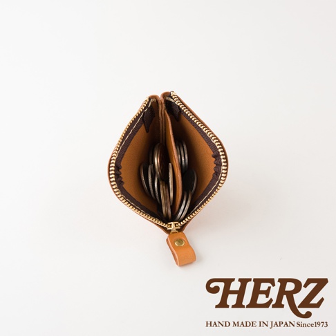 由日本HERZ職人手作的棕色皮革零錢包，呈現俯視視角，包內設有分隔層並裝有數枚硬幣，下方印有HERZ品牌商標與日本製造字樣，整體設計簡約實用。