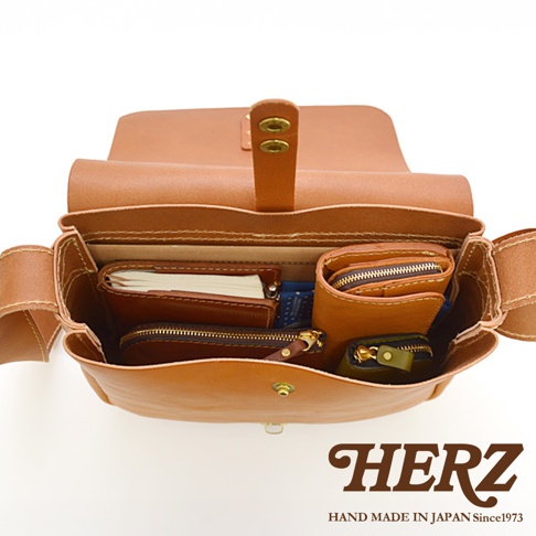 日本HERZ職人手作皮革軟皮橫式斜背包，內部收納著皮夾、筆記本等物品，展現精緻手工質感，並標有品牌HERZ及「HAND MADE IN JAPAN Since1973」。