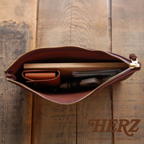 職人手作皮革Organ拉鍊式小物收納袋，展示棕色皮革材質，袋內裝有卡夾、筆記本、手機及鑰匙等小物，拉鍊為金色，整體置於木紋背景上。