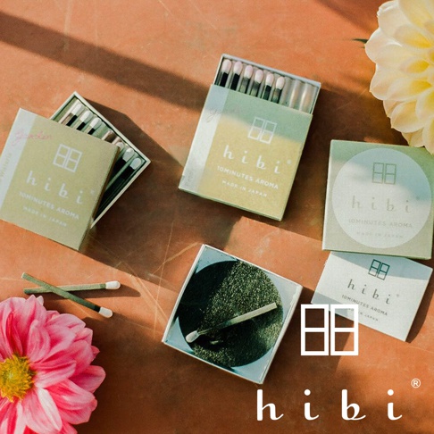 多個日式簡約風格的hibi香氛火柴盒散落在橘褐色桌面上，周圍點綴著鮮豔的粉色與淺黃色花朵，畫面呈現出柔和的自然光影與靜謐的居家香氛質感。