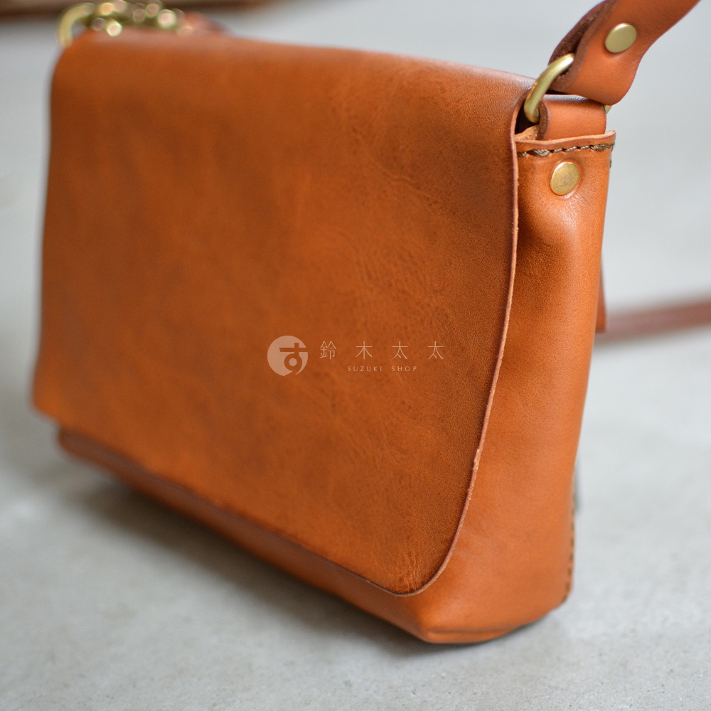 一個棕褐色的皮革斜背包，有著簡約的翻蓋設計，細膩的縫線和金屬扣環，背包的皮革表面有自然的紋理。