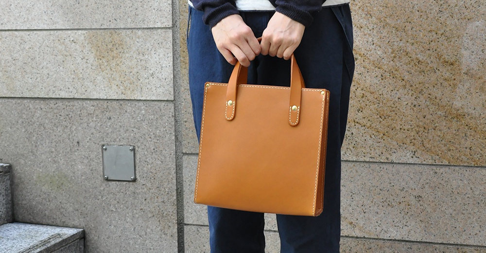 一個職人手作的橘棕色皮革直式托特包，包身簡潔，配有雙提把，以白色縫線和金色鉚釘點綴，展現皮革質感。