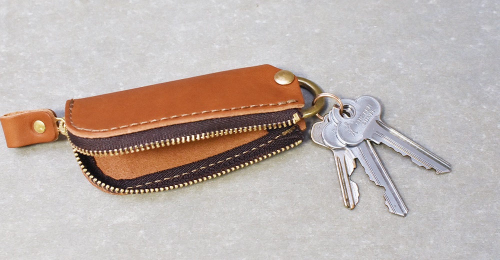 棕色皮革拉鍊鑰匙包，配有黃銅色拉鍊和金屬鑰匙圈，包包一側有一個帶有按扣的皮革提把，上面掛著幾把銀色鑰匙。