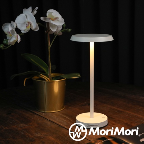 白色LED T-Light LOUNGE XL工型造型燈，頂部為圓盤形燈罩，燈身細長，底座為圓形，散發暖黃色光芒，旁邊擺放著一盆白色蝴蝶蘭，置於木質桌面。