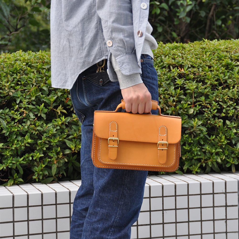 手持一個焦糖色皮革迷你肩背包，包款有雙扣設計與提把，包身縫線清晰，材質為皮革。