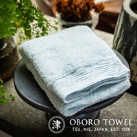 淺藍色純棉毛巾，柔軟蓬鬆，帶有細緻的條紋織邊，置於黑色陶瓷盤上，背景為綠色植物和裝飾。